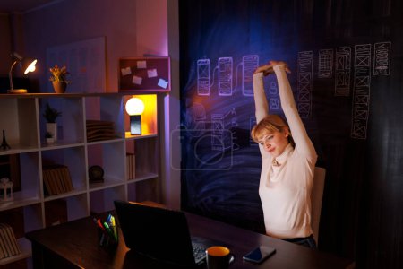 Téléchargez les photos : Femme designer UX UI faire des heures supplémentaires au bureau à la maison tard dans la nuit, s'étirant tout en étant assis à son bureau - en image libre de droit