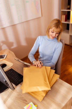 Foto de Mujer propietaria de una pequeña empresa sentada en su escritorio en la oficina en el hogar, empaquetando productos para la entrega; Empresaria mujer empaquetando pedidos en línea para envío - Imagen libre de derechos