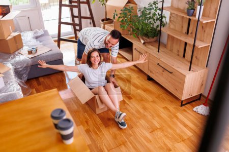 Photo for High angle view of couple in love having fun while moving in together in new home, man pushing woman around in a cardboard box - Royalty Free Image
