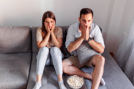 Photo for Beautiful young couple in love spending leisure time together at home, having fun watching horror movie on TV and eating popcorn - Royalty Free Image