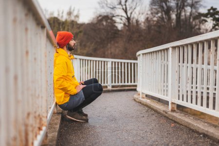 Photo for Active young man squatting and leaning at bridge fence while taking a break from jogging, doing his morning workout session - Royalty Free Image