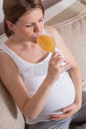 Téléchargez les photos : Femme enceinte assise sur un canapé dans un salon et buvant du jus d'orange frais - en image libre de droit