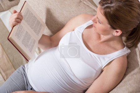 Téléchargez les photos : Belle femme enceinte assise sur un canapé dans un salon et lisant un livre - en image libre de droit