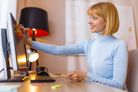 Téléchargez les photos : Femme assise à son bureau au bureau à la maison, prenant des notes tout en réfléchissant à la recherche d'idées et d'inspiration, plaçant des notes collantes sur l'écran de l'ordinateur - en image libre de droit