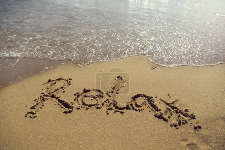 Téléchargez les photos : Word relax écrit à la main dans du sable doré sur la plage à côté de la ligne de flottaison - en image libre de droit