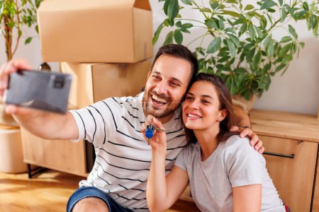 Téléchargez les photos : Jeune couple amoureux emménageant ensemble dans leur nouvel appartement, s'amusant à prendre des selfies avec les clés de l'appartement - en image libre de droit