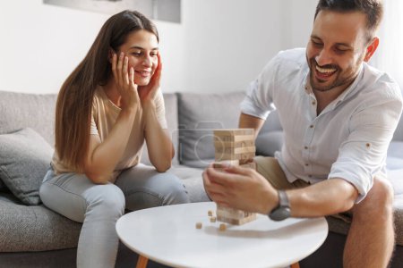 Téléchargez les photos : Joyeux jeune couple s'amuser passer du temps libre ensemble à la maison en jouant bloc empilement et enlever jeu d'équilibrage - en image libre de droit