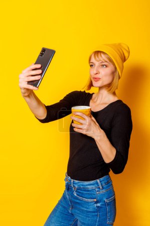 Foto de Retrato de una hermosa mujer rubia usando sombrero tomando una selfie mientras bebe café sobre fondo de color amarillo - Imagen libre de derechos