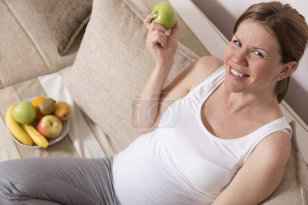 Téléchargez les photos : Belle femme enceinte assise sur un canapé dans un salon avec un bol de fruits placé à côté d'elle, mangeant une pomme - en image libre de droit