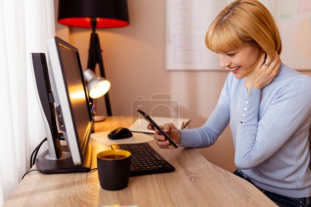 Téléchargez les photos : Femme assise à son bureau à la maison, tapant un message texte à l'aide d'un téléphone intelligent tout en travaillant - en image libre de droit