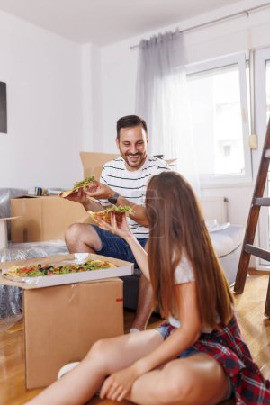 Photo for Couple in love moving in together, taking a break from unpacking cardboard boxes, having fun while eating pizza for lunch - Royalty Free Image