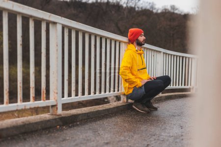 Photo for Active young man squatting and leaning at bridge fence while taking a break from a morning run - Royalty Free Image