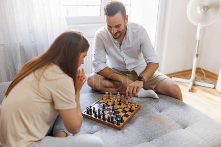 Téléchargez les photos : Couple amoureux assis sur le canapé dans le salon, s'amuser à jouer aux échecs et profiter du temps libre à la maison - en image libre de droit