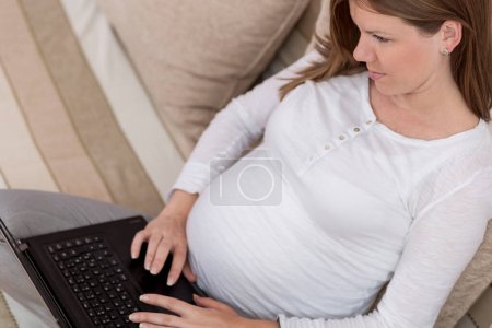 Foto de Mujer embarazada sentada en un sofá en una sala de estar y navegando por la red en una computadora portátil - Imagen libre de derechos