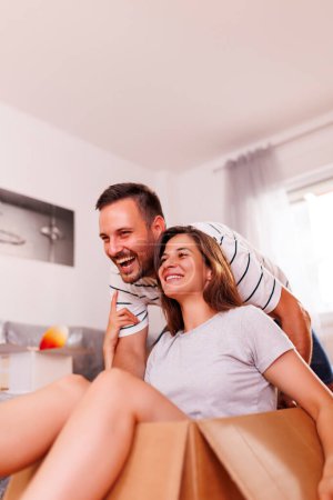 Photo for Young couple in love moving in together, having fun while unpacking cardboard boxes with their belongings, boyfriend pushing girlfriend around in a cardboard box - Royalty Free Image