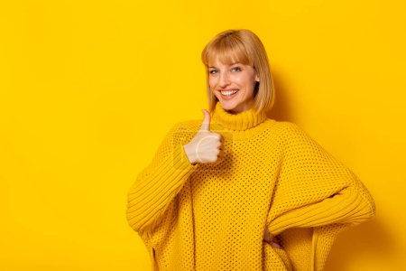 Foto de Hermosa mujer rubia en suéter amarillo sonriendo y mostrando los pulgares hacia arriba en el fondo de color amarillo con espacio de copia - Imagen libre de derechos