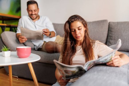 Téléchargez les photos : Beau couple lisant le journal et buvant du café tout en passant du temps libre ensemble à la maison le matin - en image libre de droit