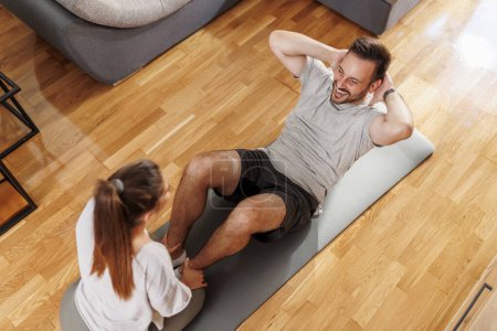 Photo for High angle view of couple working out together at home, man doing crunches while woman holding his legs and counting - Royalty Free Image