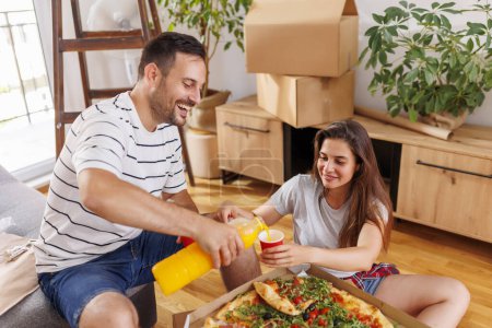 Téléchargez les photos : Beau couple amoureux prenant une pause tout en déménageant dans une nouvelle maison ensemble, boire du jus, manger de la pizza et se détendre - en image libre de droit