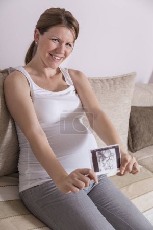 Téléchargez les photos : Femme enceinte assise sur un canapé dans le salon d'un appartement, tenant et regardant une photo échographique de son bébé - en image libre de droit