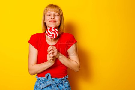 Téléchargez les photos : Belle femme en vêtements d'été tendance tenant des bonbons rouges et blancs avec les yeux fermés sur fond jaune avec espace de copie - en image libre de droit