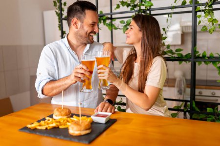 Photo for Beautiful young couple in love having fun spending leisure time together at home, making a toast while eating burgers and drinking beer - Royalty Free Image
