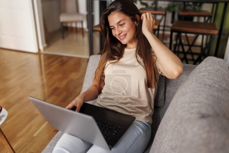 Téléchargez les photos : Belle jeune femme allongée sur le canapé avec un ordinateur portable sur ses genoux, riant tout en ayant un appel vidéo avec des amis - en image libre de droit