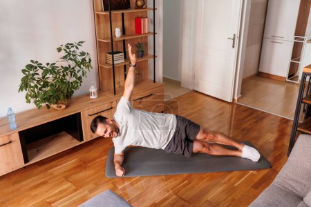 Foto de Hombre activo guapo haciendo ejercicio en casa, haciendo tablón lateral en estera de yoga; hombre haciendo ejercicio en casa por la mañana - Imagen libre de derechos