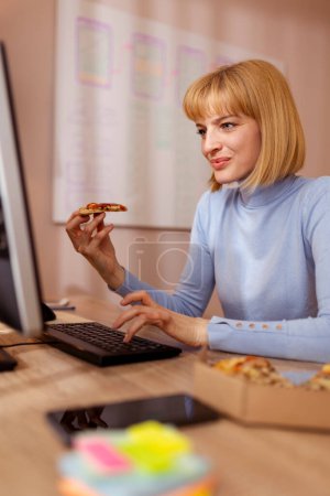 Téléchargez les photos : Femme assise à son bureau à la maison, mangeant de la pizza tout en travaillant - en image libre de droit