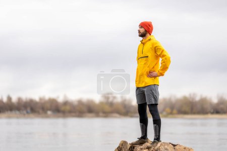 Téléchargez les photos : Homme debout au bord de la rivière sur un tronc d'arbre tombé, faisant une pause en faisant du jogging un jour d'automne couvert - en image libre de droit