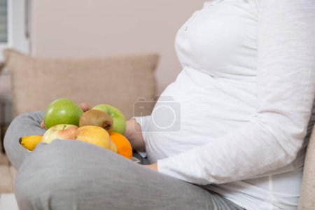 Téléchargez les photos : Gros plan du ventre d'une femme enceinte alors qu'elle est assise sur un canapé dans un salon, et tenant un bol de fruits - en image libre de droit