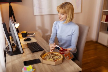Téléchargez les photos : Femme assise à son bureau tout en travaillant au bureau à la maison, manger de la pizza tout en prenant une pause déjeuner - en image libre de droit