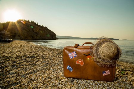 Téléchargez les photos : Valise vintage avec autocollants drapeau et chapeau de paille, placée sur la plage avec mer paisible au lever du soleil sur l'île de Thassos en Grèce - en image libre de droit