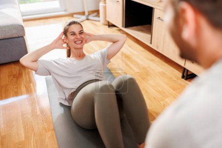 Photo for Couple working out together at home, woman lying on yoga mat doing sit ups while man is holding her legs and counting - Royalty Free Image