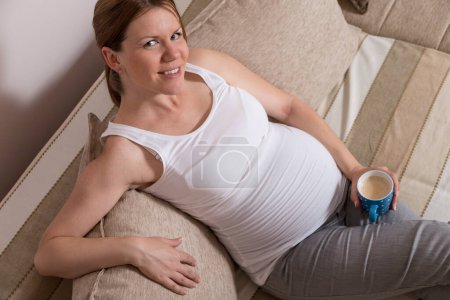 Téléchargez les photos : Femme enceinte assise sur un canapé dans un salon, tenant une tasse de café - en image libre de droit