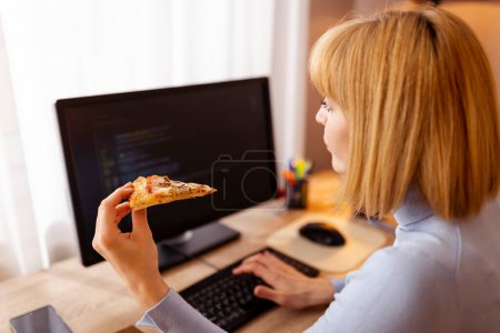 Foto de Desarrolladora de software femenina trabajando en la oficina en casa, sentada en su escritorio y comiendo pizza mientras codifica - Imagen libre de derechos