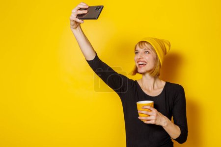 Foto de Retrato de una hermosa mujer rubia usando sombrero tomando una selfie mientras bebe café sobre fondo de color amarillo con espacio de copia - Imagen libre de derechos