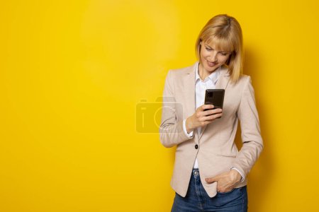 Téléchargez les photos : Belle confiante jeune femme d'affaires dactylographier un message texte à l'aide d'un téléphone intelligent isolé sur fond jaune avec espace de copie - en image libre de droit
