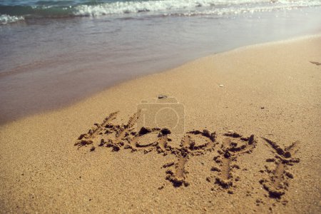 Téléchargez les photos : Mot heureux écrit à la main dans le sable sur la plage à côté de la ligne de flottaison - en image libre de droit