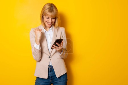 Jeune femme excitée gagnant pari en ligne en utilisant l'application de pari de téléphone intelligent isolé sur fond jaune avec espace de copie