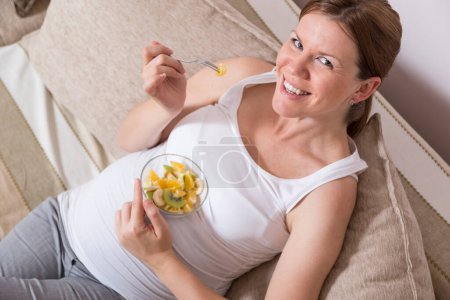 Téléchargez les photos : Femme enceinte assise sur un canapé dans un salon et mangeant une salade de fruits - en image libre de droit