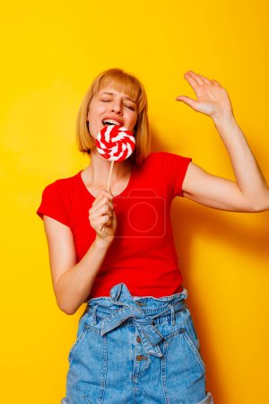 Téléchargez les photos : Belle femme blonde en tenue d'été tendance tenant des bonbons comme micro chantant et dansant sur fond de couleur jaune - en image libre de droit