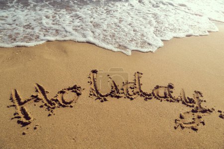 Téléchargez les photos : Mot vacances écrit à la main dans le sable doré sur la plage à côté de la ligne de flottaison - en image libre de droit