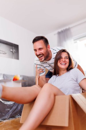 Photo for Beautiful couple in love moving in new home, having fun while unpacking cardboard boxes with their belongings, husband pushing wife around in a cardboard box - Royalty Free Image