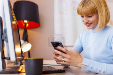Photo for Woman sitting at her desk in home office, working remotely from home, holding smart phone and taking notes in planner - Royalty Free Image
