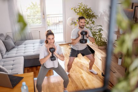 Photo for Couple working out at home doing squats using dumbbells - Royalty Free Image