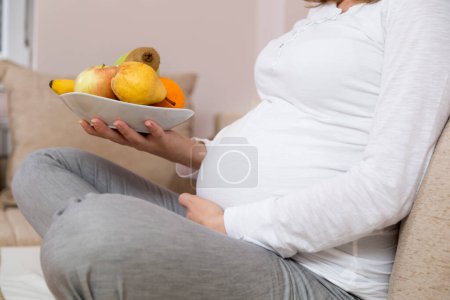 Téléchargez les photos : Gros plan du ventre d'une femme enceinte alors qu'elle est assise sur un canapé dans un salon, et tenant un bol de fruits - en image libre de droit