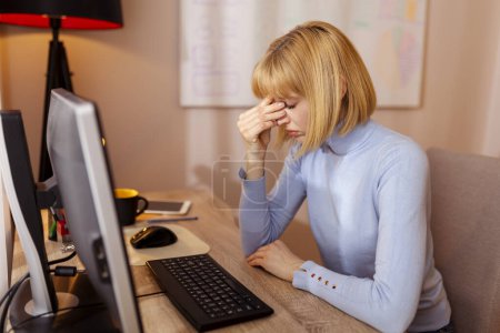 Frau arbeitet in einem Büro, sitzt an ihrem Schreibtisch, hält den Kopf in den Händen, hat Kopfschmerzen