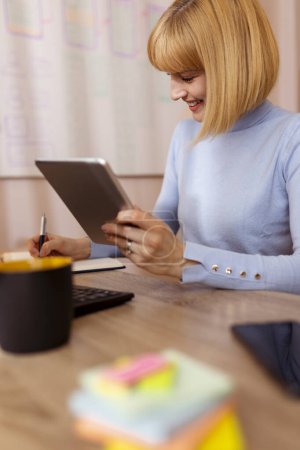 Téléchargez les photos : Femme assise à son bureau à la maison, suivant un cours en ligne à l'aide d'un ordinateur tablette, écrivant dans un planificateur - en image libre de droit