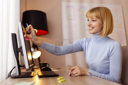 Téléchargez les photos : Femme assise à son bureau au bureau à la maison, prenant des notes tout en réfléchissant à la recherche d'idées et d'inspiration, plaçant des notes collantes sur l'écran de l'ordinateur - en image libre de droit